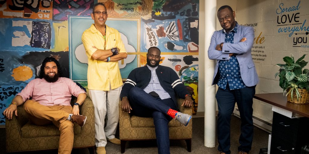 4 men sitting and standing around a couch posing for the camera